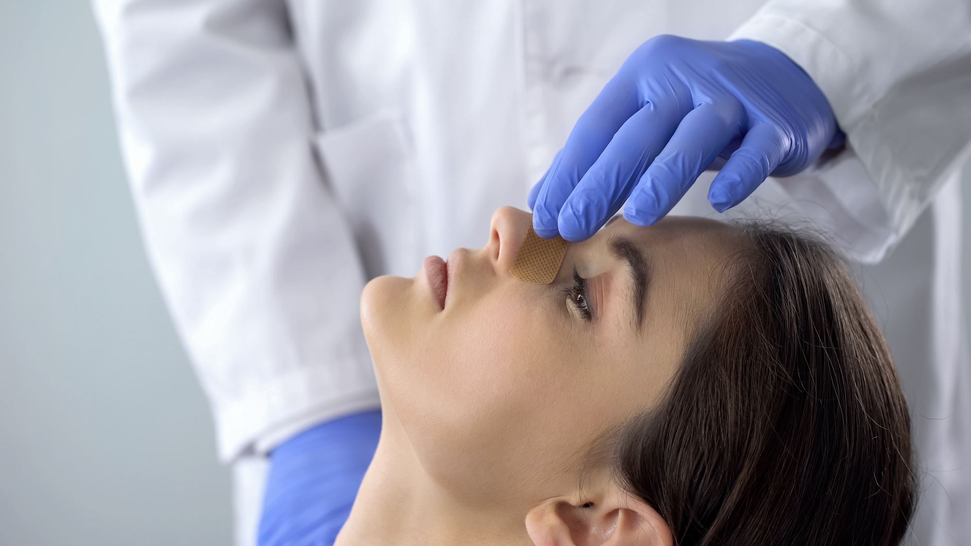 person in white coat with blue gloves with hands on young woman's nose