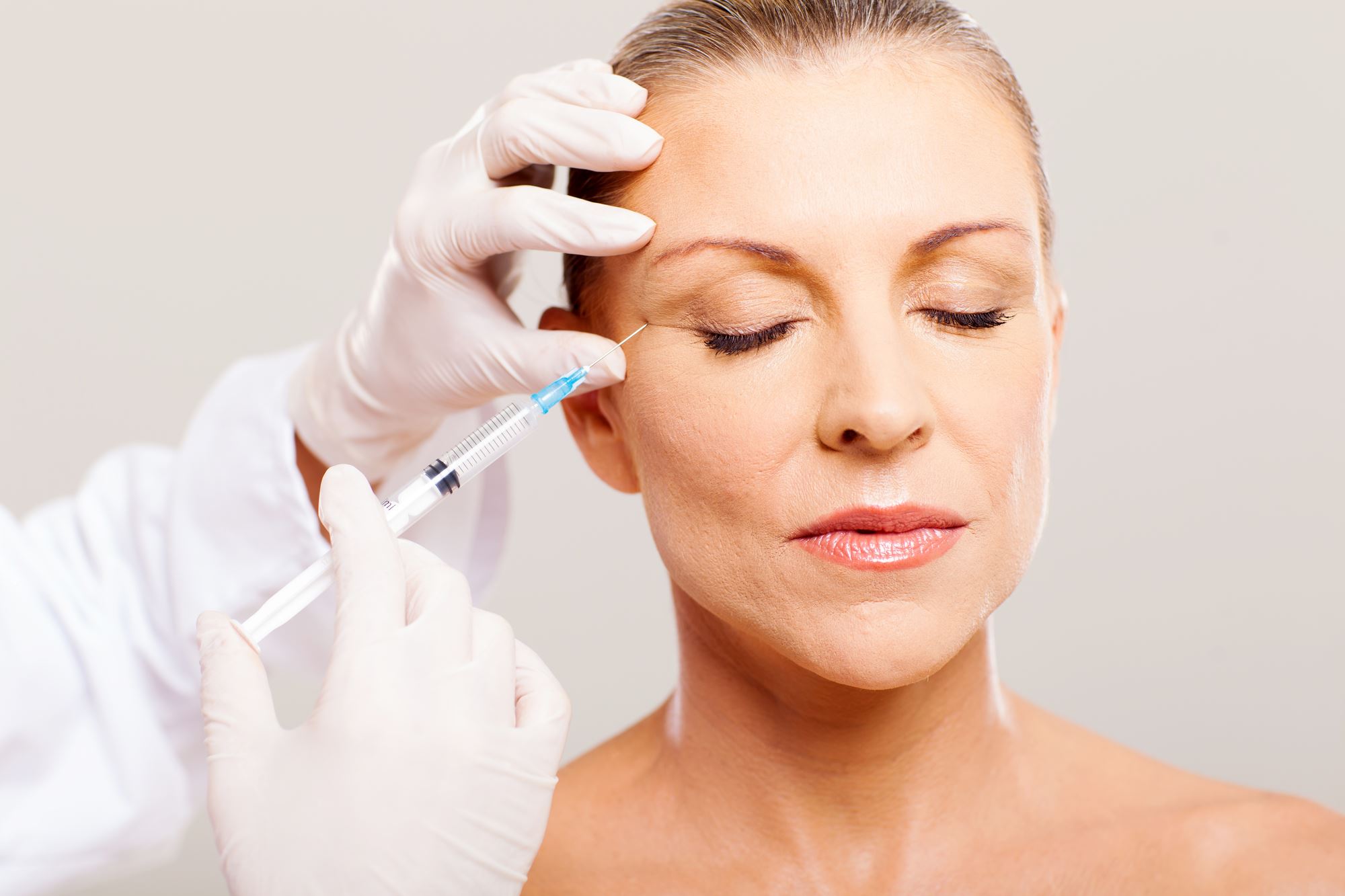 doctor wearing white coat and gloves holding syringe injecting botox into mature woman eyebrow