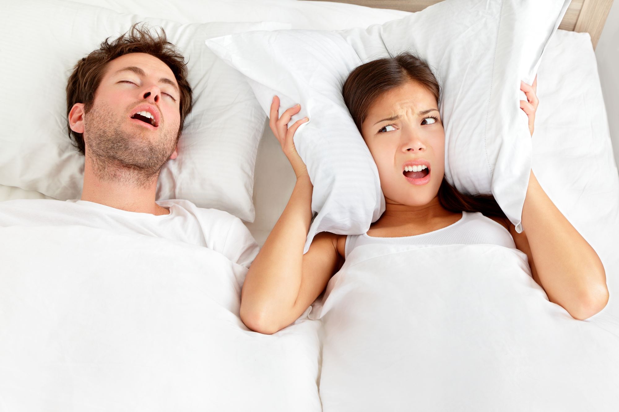 young couple lying on white bed with man snoring and woman holding pillow around ears