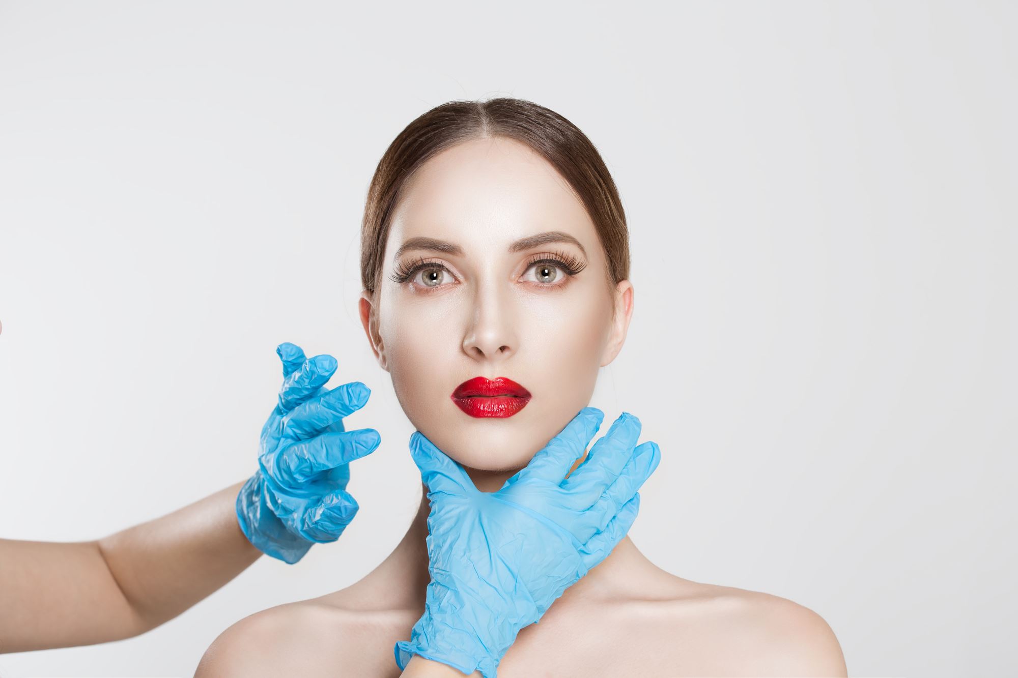 young woman wearing red lipstick on white background with blue gloved doctor hands around chin