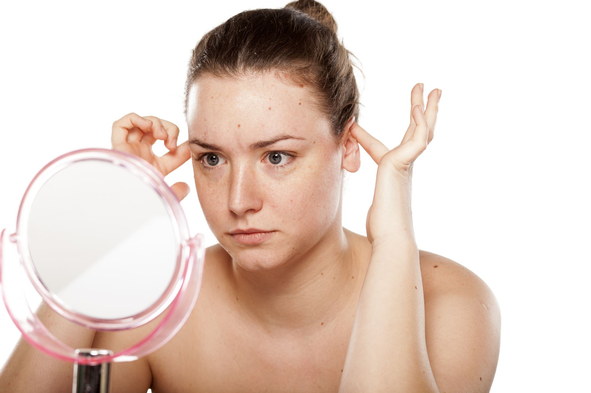 Young brown haired woman with hair pulled back looking into mirror holding ears considering otoplasty
